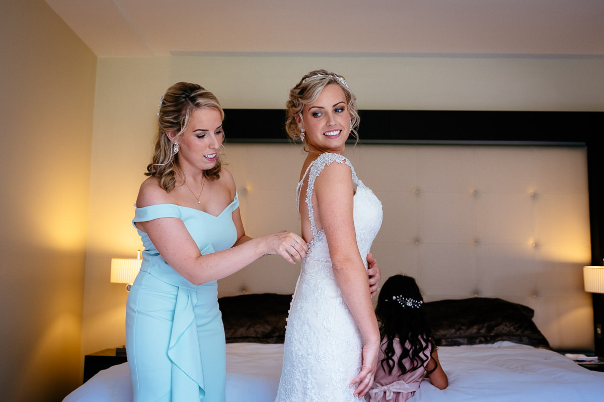 A bridesmaid helps fasten the back of the bride's dress as a flower girl looks on at Carton House.