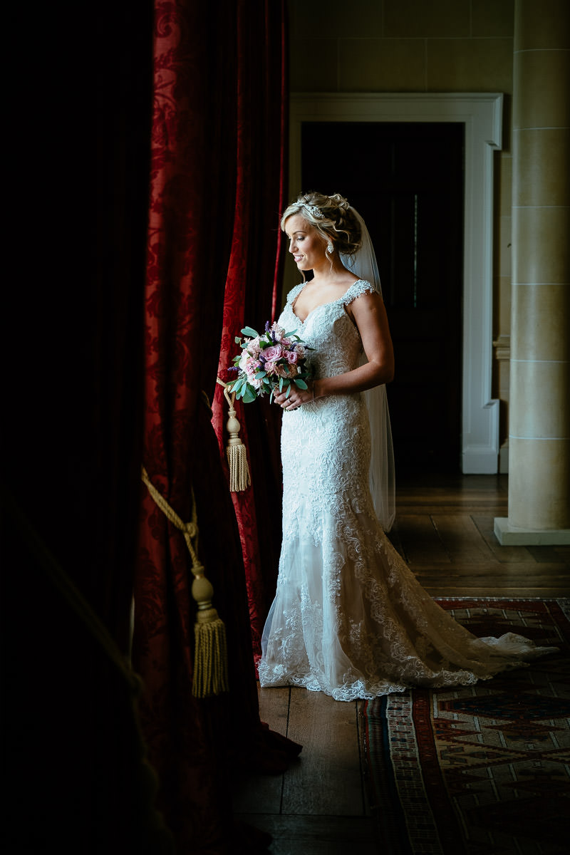 The bride stands by a red curtain holding her bouquet before the ceremony at Carton House.