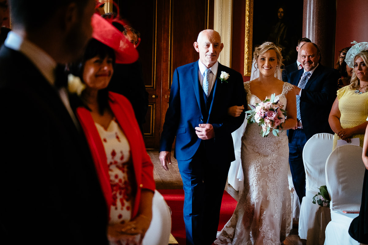 The bride walks down the aisle with her father past smiling guests at Carton House.