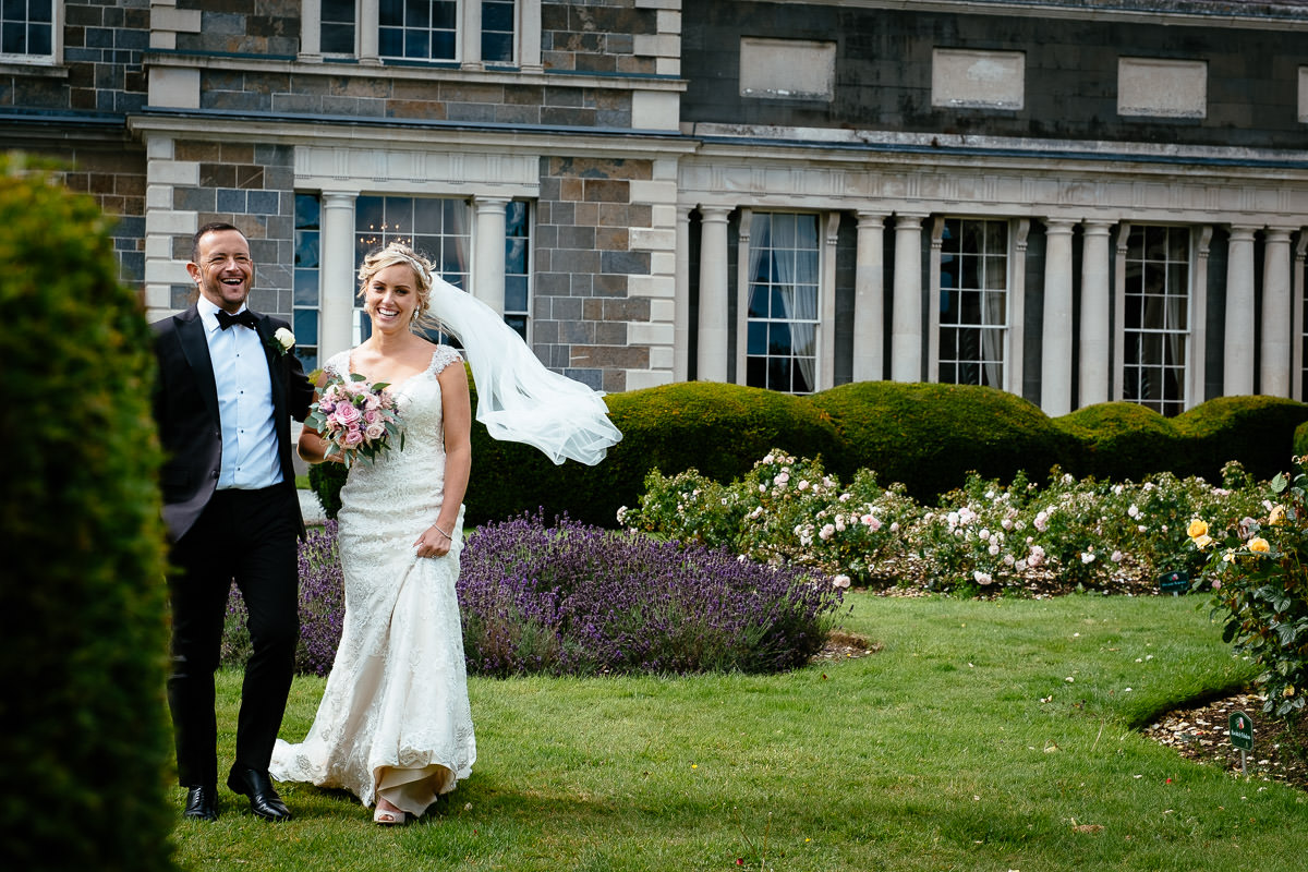 The bride and groom walk together across the lawn as her veil blows in the wind at Carton House.