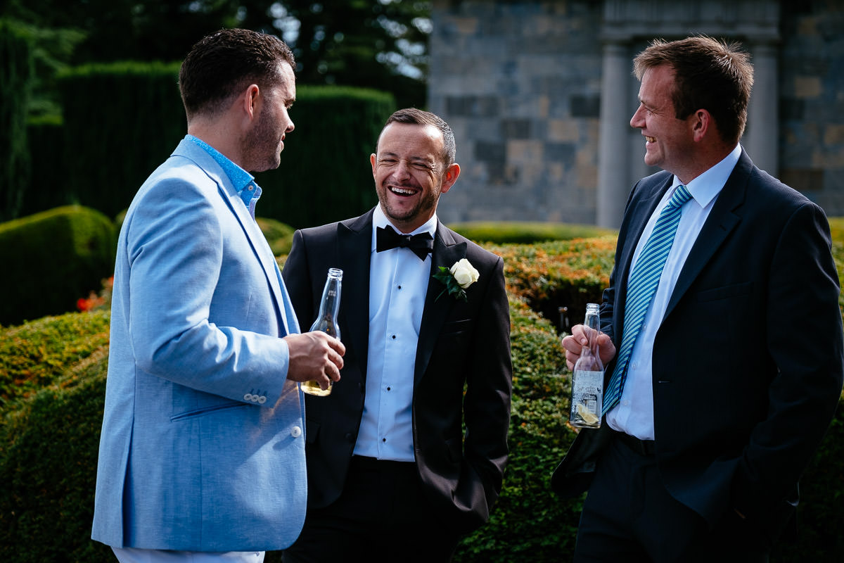 The groom laughs with two guests holding bottles of beer in the gardens at Carton House.