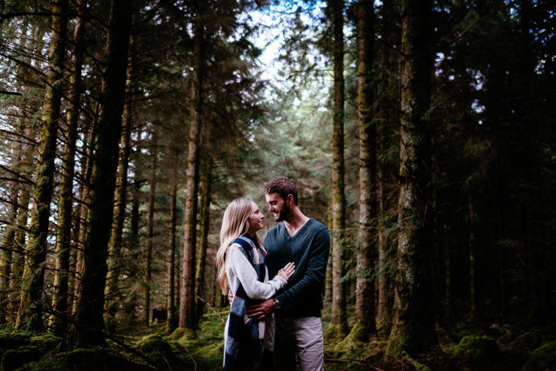 A man and woman stand close together, looking at each other during an engagement photoshoot in a dense forest in rural Ireland.