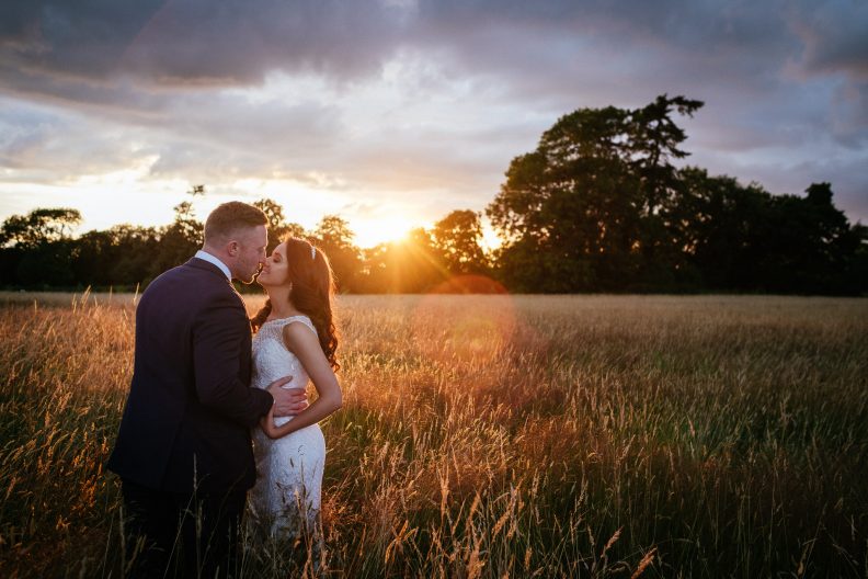 bride and groom kissing in a field of wheat with the sun setting behind them
