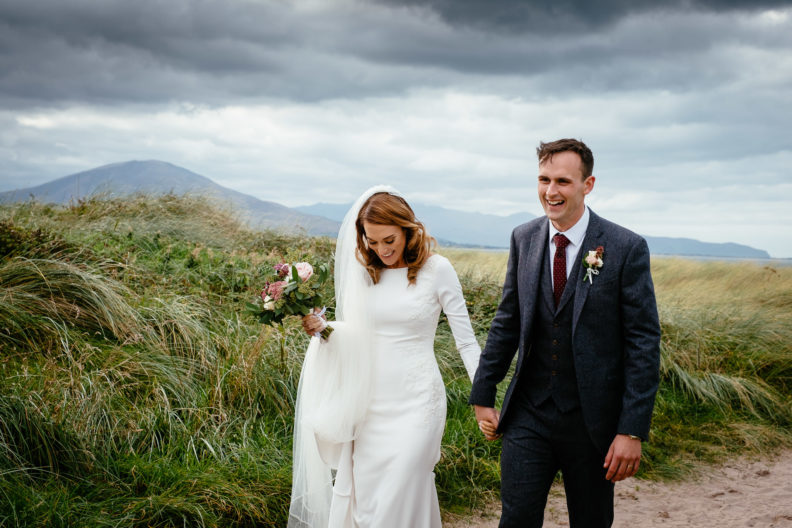 Beautiful Ballyseede Castle Wedding 8 bride and groom laughing and walking along beach in west of ireland with mountains in background