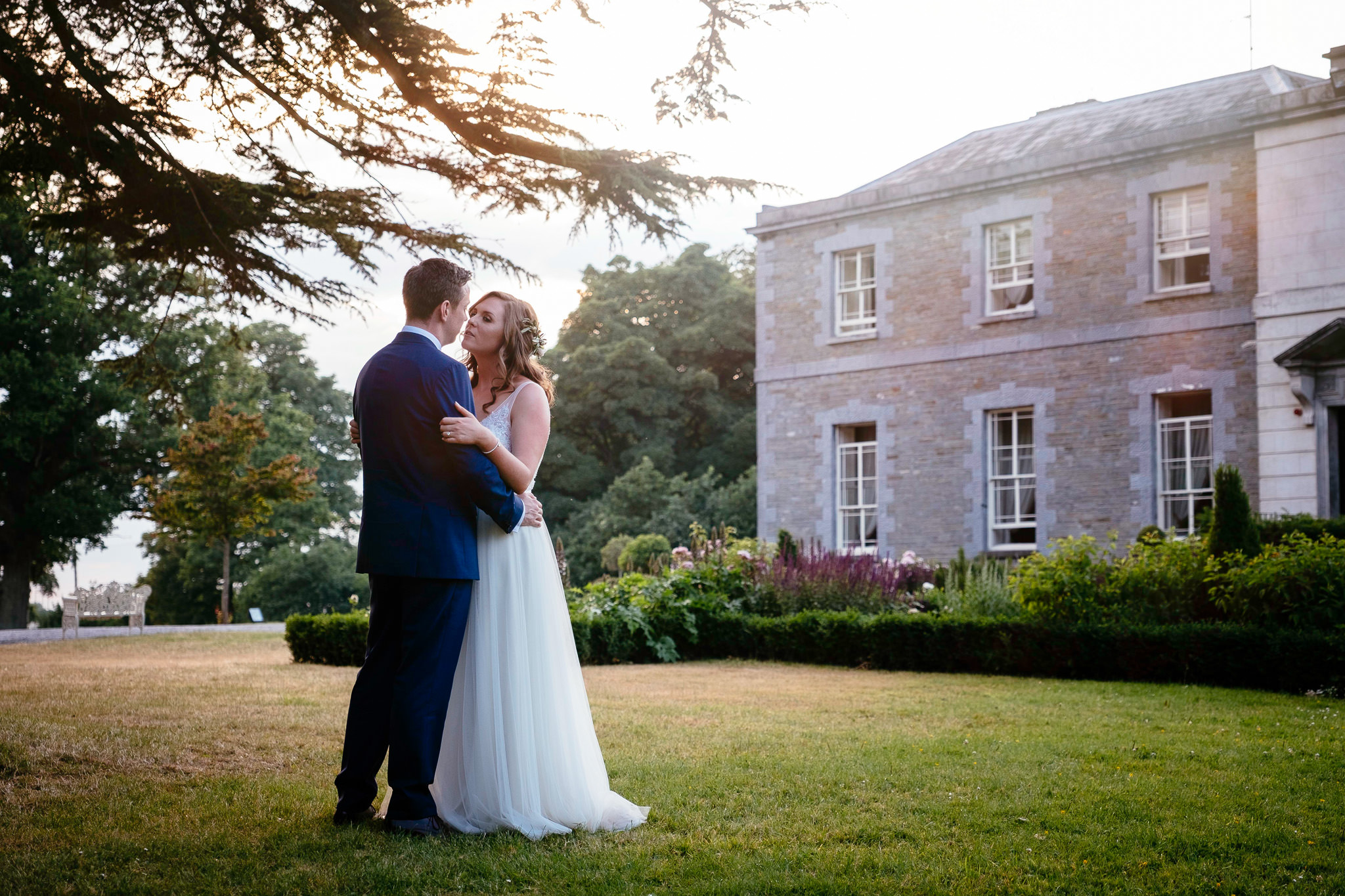 A bride and groom embrace and share a kiss on the lawn near the grand stone walls of Tankardstown House at sunset.