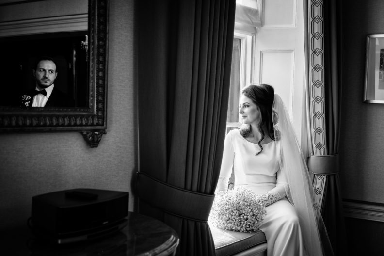 Bride sitting by a window with her grooms reflection in a mirror