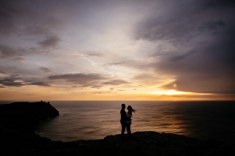 engaged couple at the Cliffs of Moher Ireland with the sun setting over the ocean