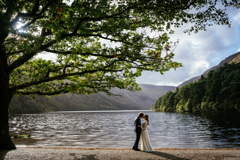 A bride and groom stand forehead-to-forehead on a lakeside shore, framed by a large tree with mountains in the background, making this the best location in Ireland for an elopement.