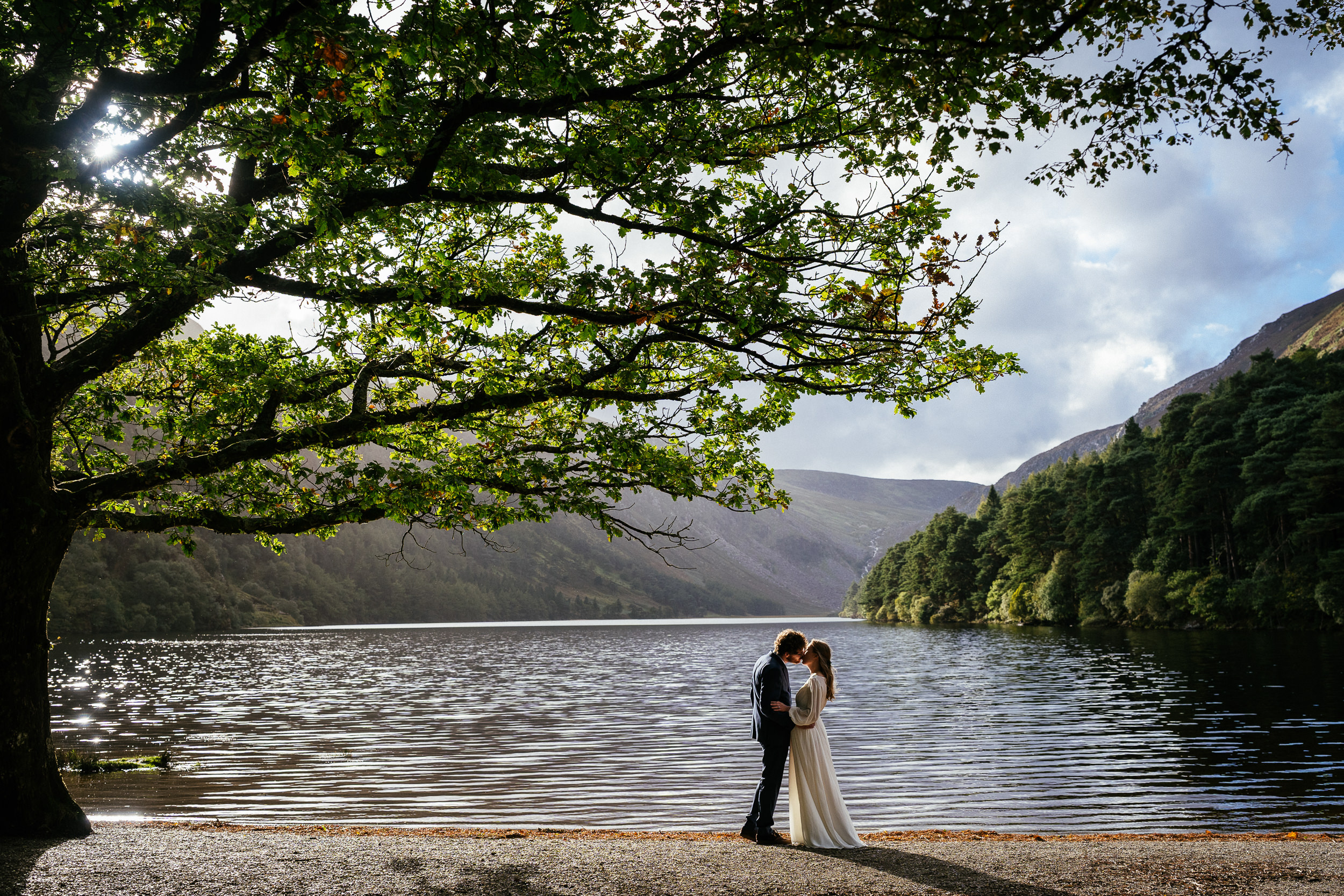 A bride and groom stand forehead-to-forehead on a lakeside shore, framed by a large tree with mountains in the background, making this the best location in Ireland for an elopement.