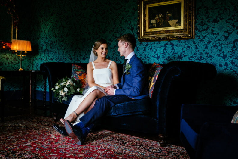 The bride and groom sit closely together on a velvet sofa in a richly decorated room at Boyne Hill House.