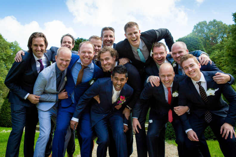 Wedding Gallery 23 A group of men in suits, smiling and posing closely together outdoors, with trees and a blue sky in the background.