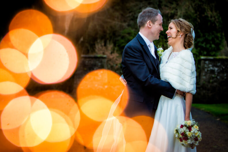 Wedding Gallery 24 A couple in wedding attire stands outdoors, smiling at each other. The bride holds a bouquet, and bokeh lights illuminate the scene.