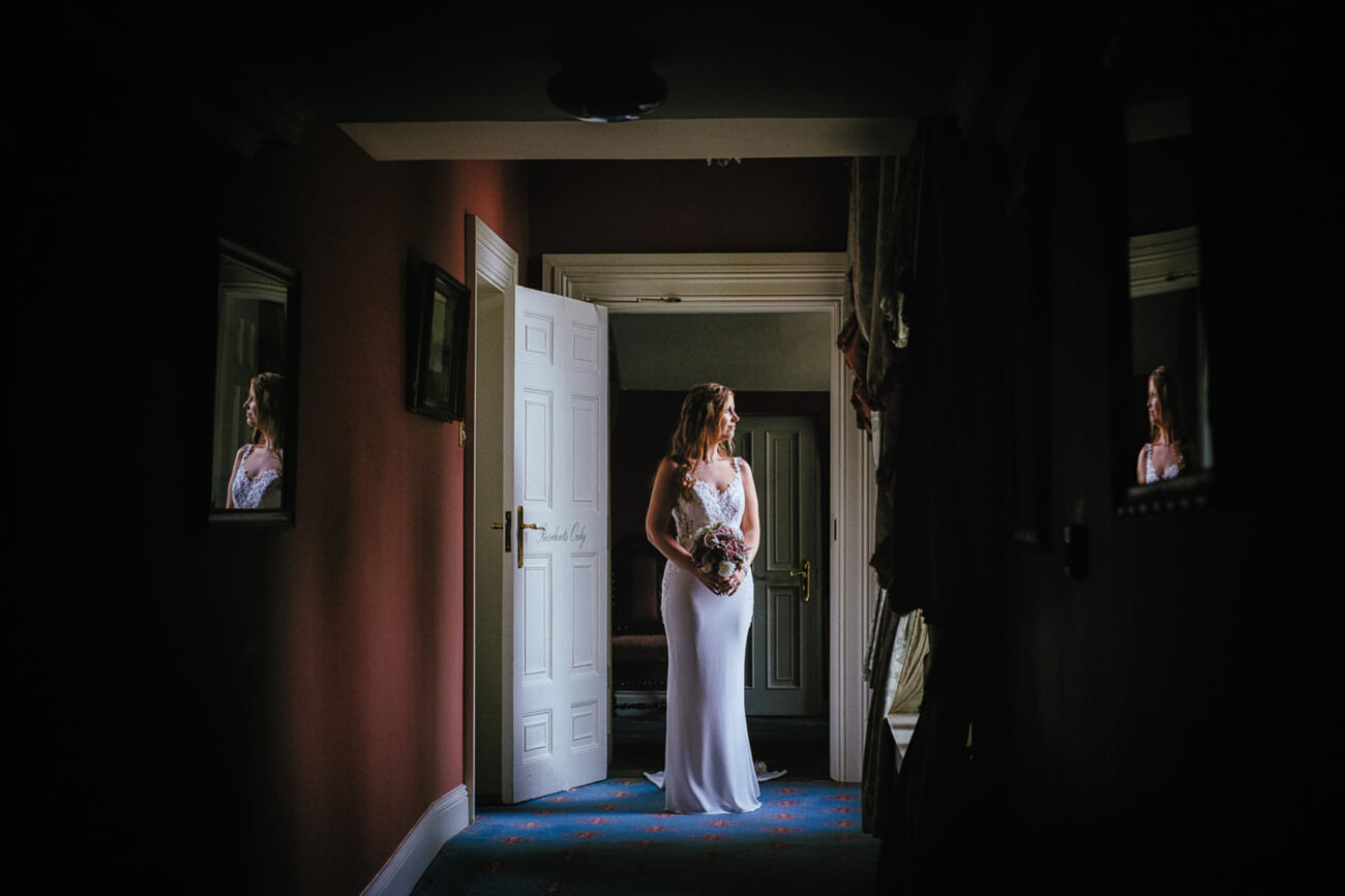 A bride in a white dress holds a bouquet, standing in a dimly lit room with a door open behind her and mirrors on the walls.