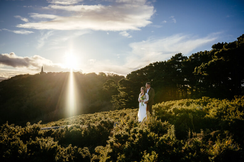 A couple in wedding attire stands on a hillside with greenery, silhouetted against the setting sun and a partly cloudy sky.