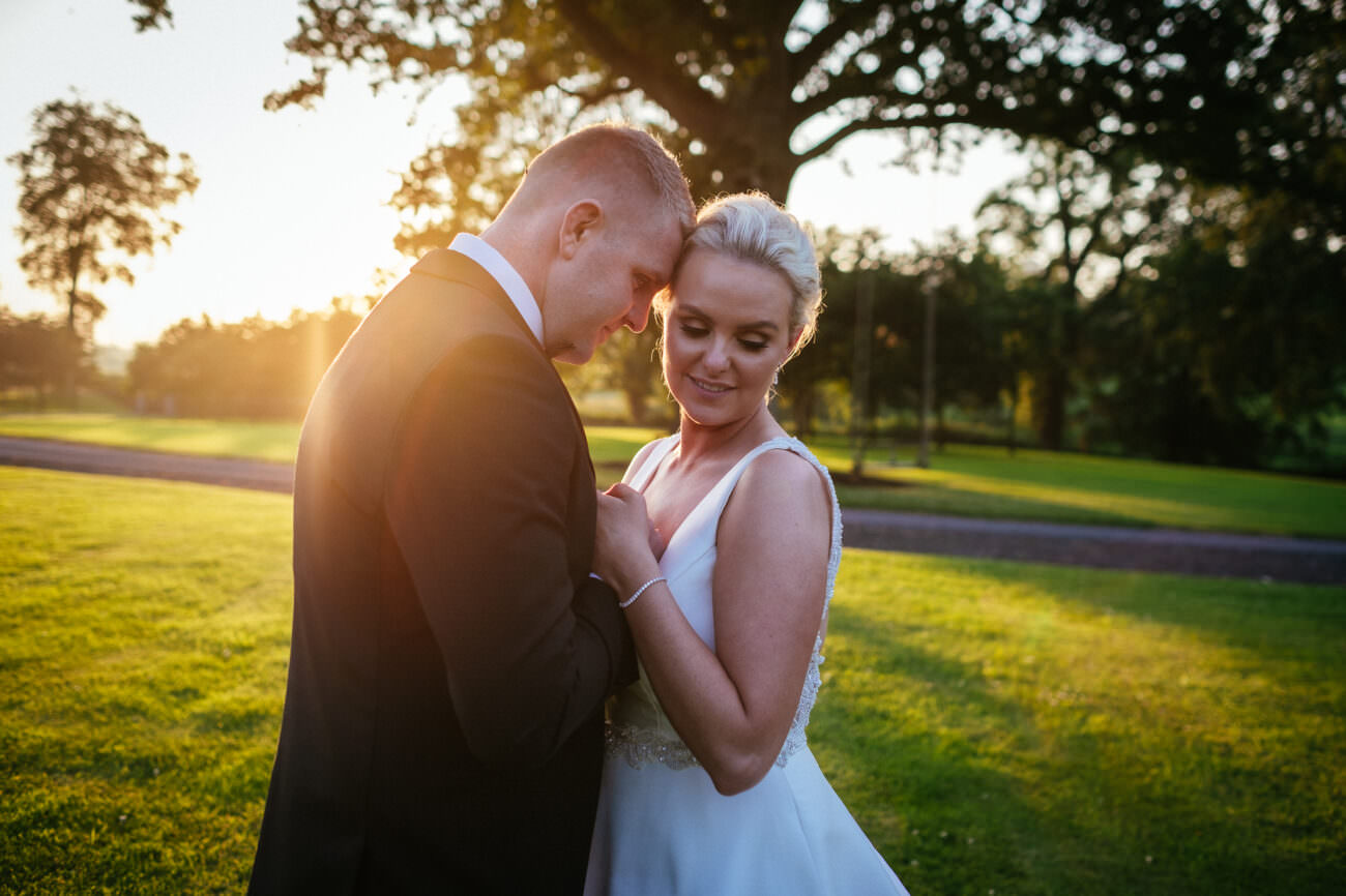 A bride and groom stand close together on a grassy lawn at sunset, holding hands and facing each other in the grounds of Tankardstown House at sunset.