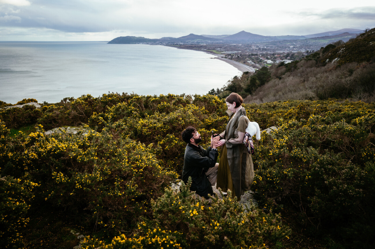 A person kneels and proposes to another during an engagement photoshoot on Killiney hill Dublin Ireland covered with greenery, overlooking a breathtaking coastal landscape with mountains and the sea in the background.
