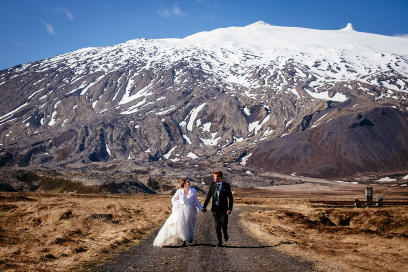 bride & groom running down a grit road with a snow capped mountain behind them on the Snæfellsnes Peninsula during her elopement