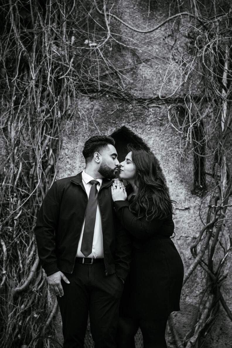 A black and white photo of a couple kissing in front of a stone wall.