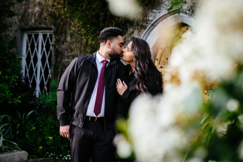 An engaged couple kissing in front of a stone wall.