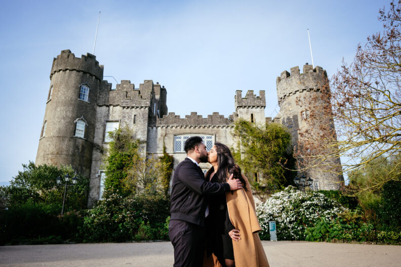 An engaged couple kissing in front of a castle.