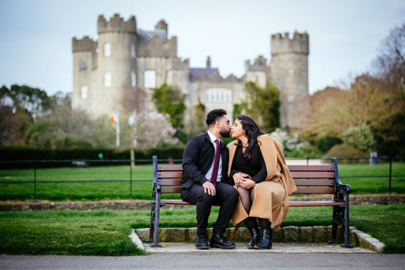 A couple kissing on a bench in front of a castle.