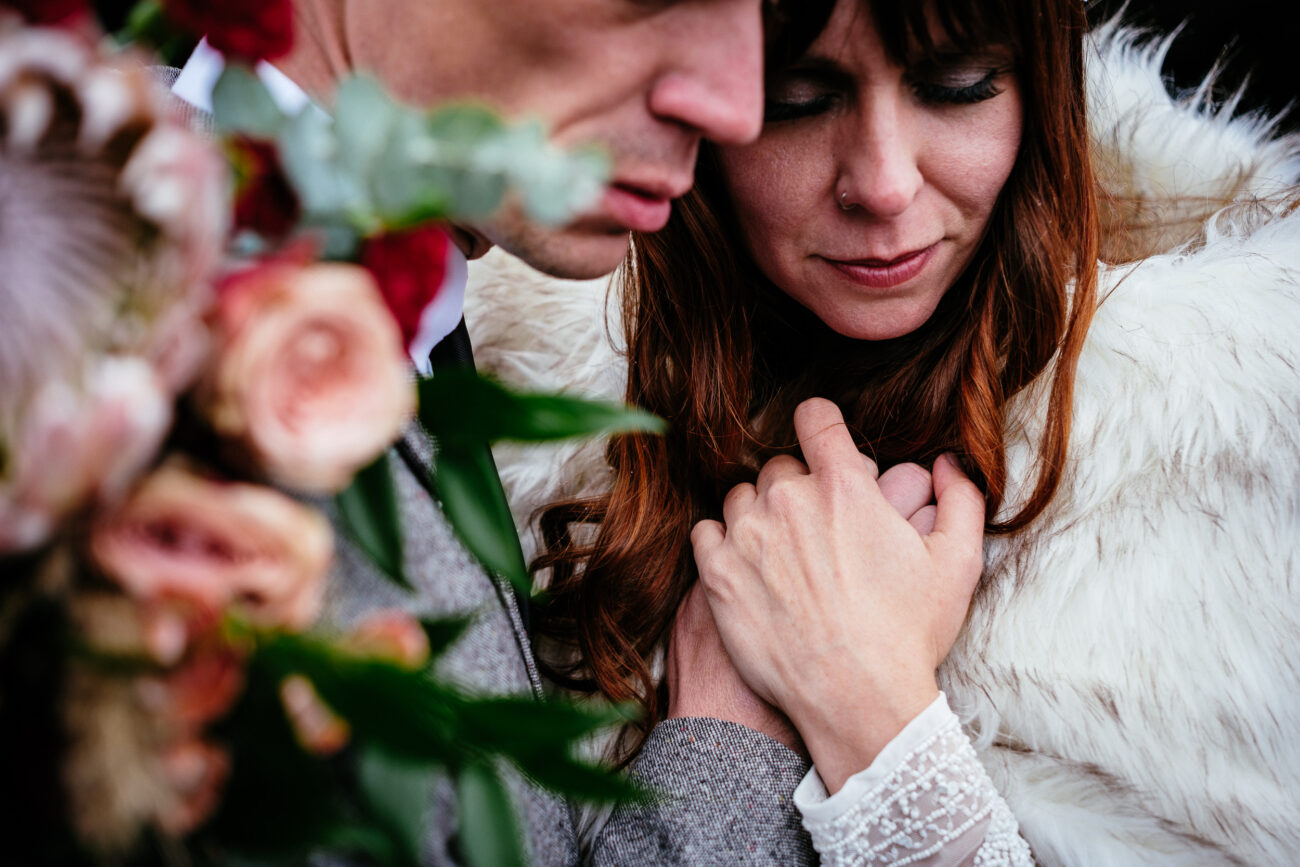 Close-up of a couple embracing, with the woman's hand gently holding the man's chest. The woman is wearing a white fur garment, and the man is in a gray suit. A bouquet of flowers is visible in the foreground.