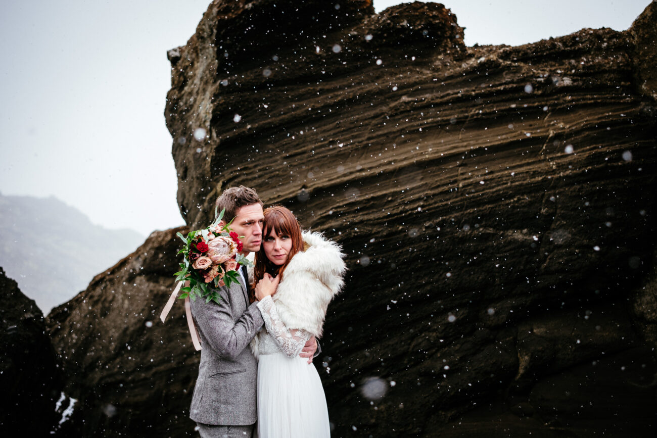 A couple in wedding attire stands close together in front of rocky terrain, with snowflakes falling around them. The groom holds a bouquet, and the bride wears a white fur stole.