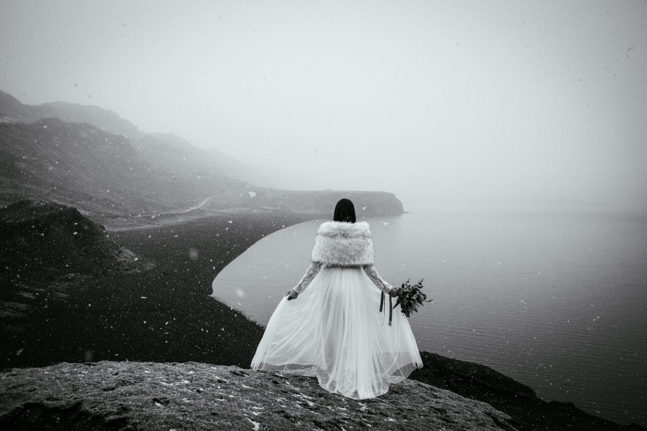 A bride in a white dress and fur wrap stands on a rocky cliff, overlooking a foggy, snow-dusted landscape by the sea, holding a bouquet in her left hand.