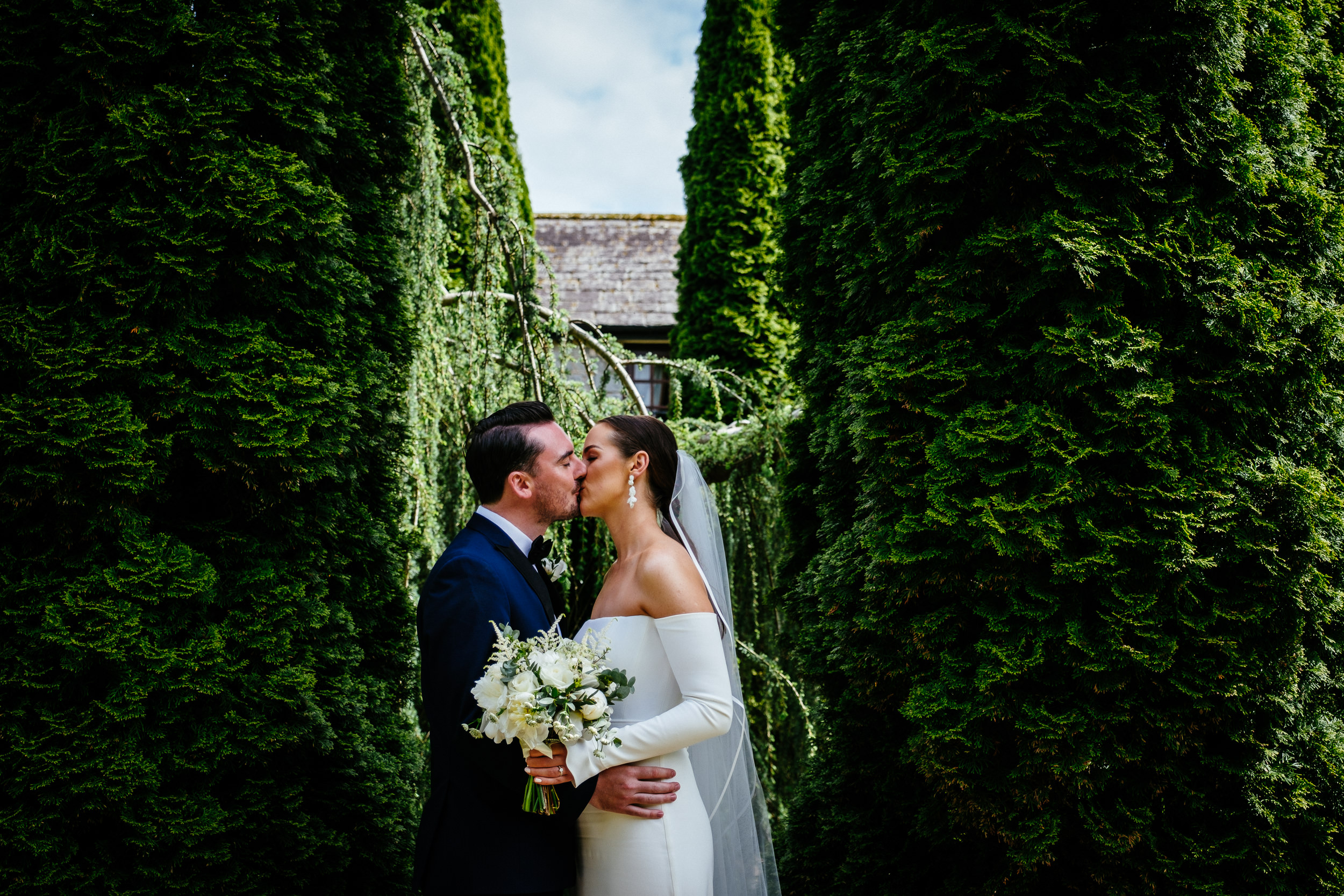 A bride and groom kiss amidst tall, lush green hedges under a clear sky at their Cabra Castle wedding.