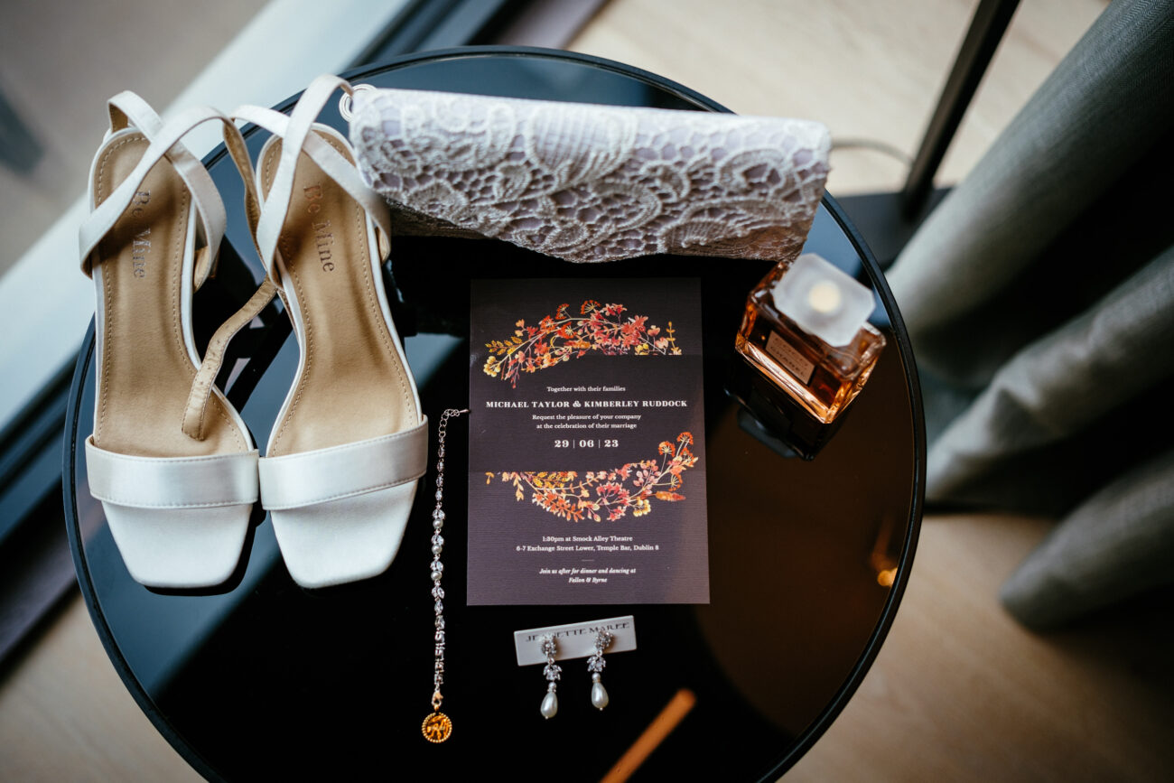 Black table displaying wedding accessories including white high-heeled shoes, lace clutch, floral wedding invitation, perfume, bracelet, and earrings.