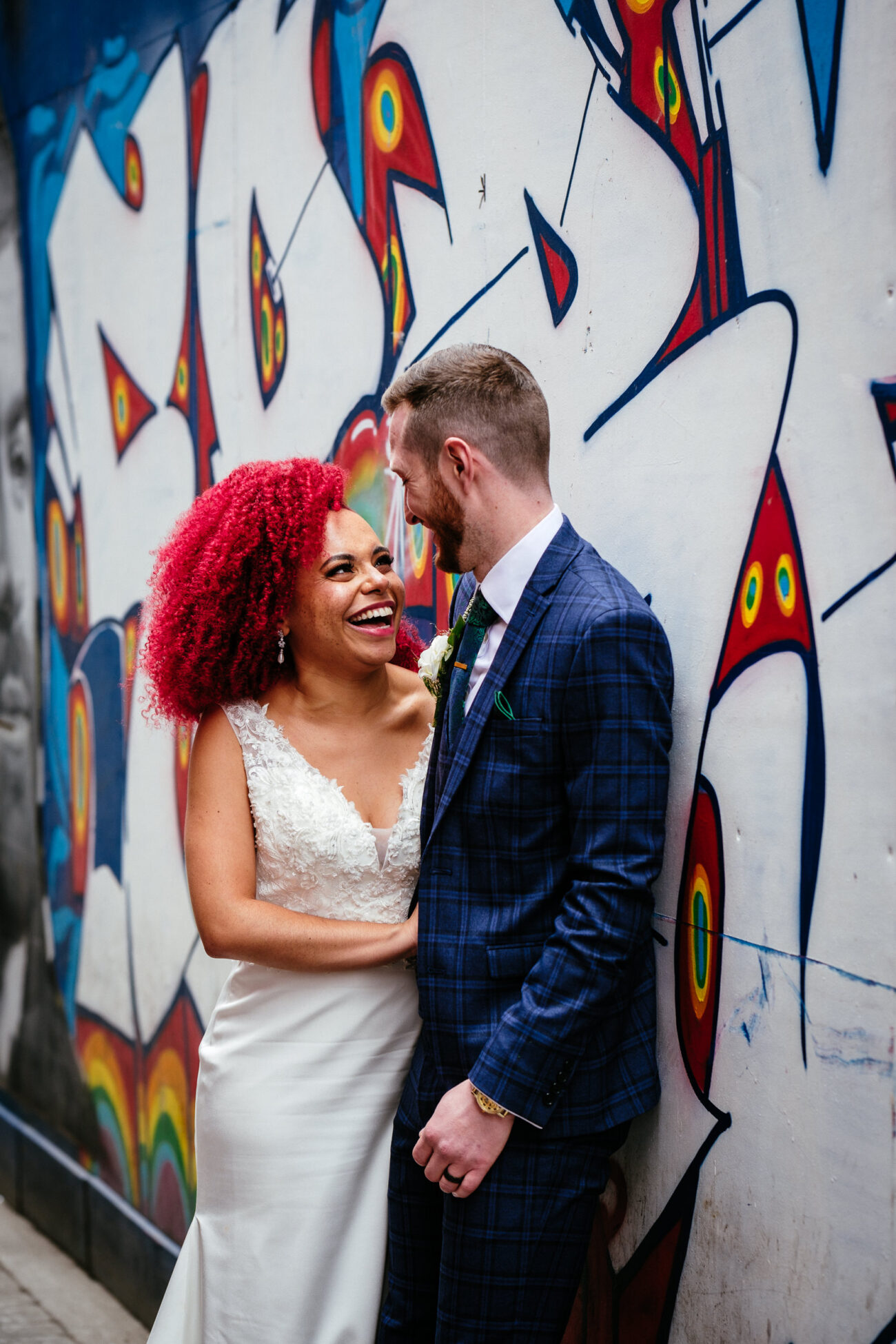A bride in a white dress and a groom in a blue suit share a joyful moment in front of a colorful graffiti wall.