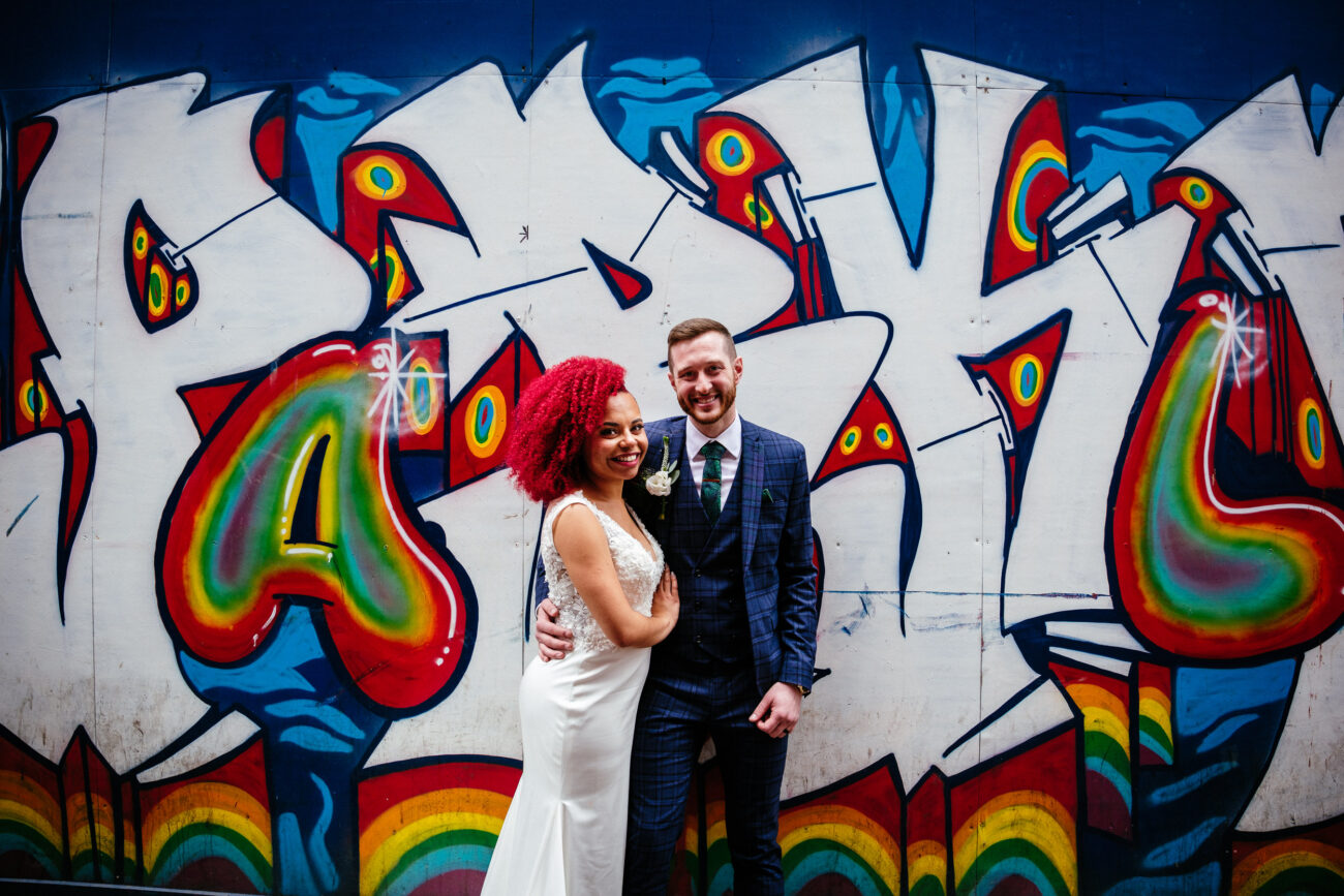 A couple in formal attire stands in front of a colorful graffiti mural with rainbow accents, capturing the joyful essence of their Smock Alley wedding. They smile at the camera, with the woman wearing a white dress and the man in a dark suit.