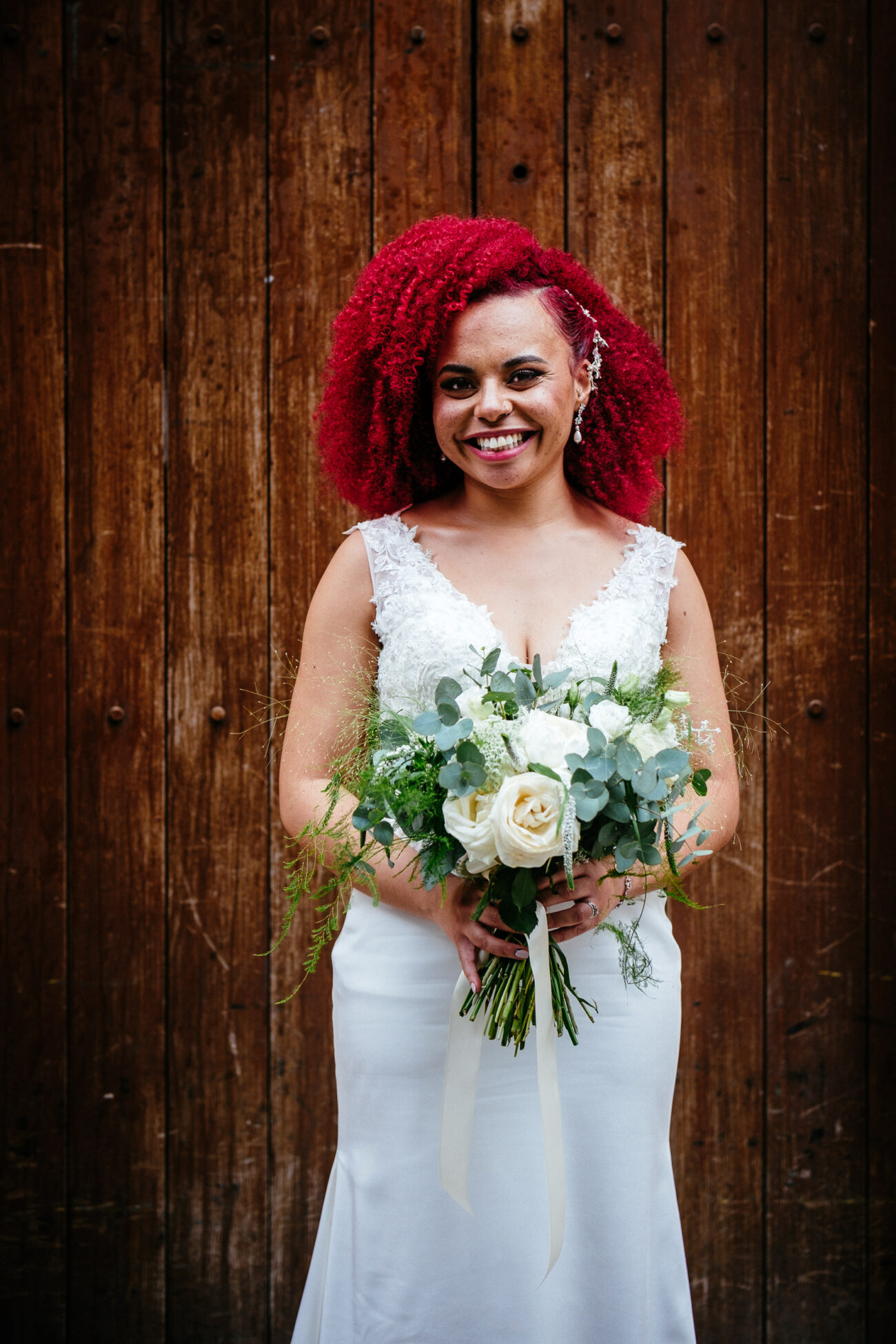 A woman with bright red hair in a white wedding dress holds a bouquet of flowers and stands in front of a wooden wall.