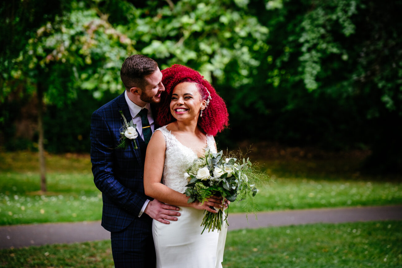 A bride holding a bouquet smiles as the groom, in a blue checkered suit, stands behind her with hands on her waist in a green outdoor setting.