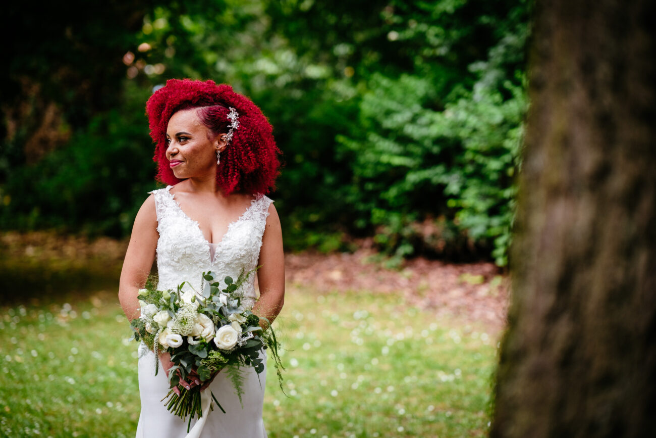 A bride with bright red hair stands outdoors in front of greenery, holding a bouquet of white flowers. She is wearing a white lace wedding dress and looking to her left.