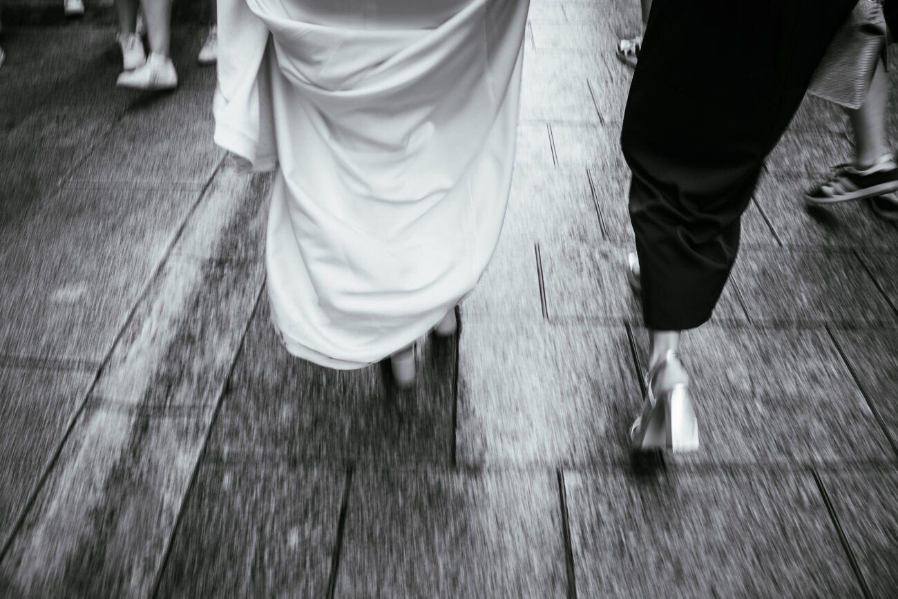 Black and white image capturing the lower half of two people walking on a stone pavement; one wears a long white dress and the other wears black pants.