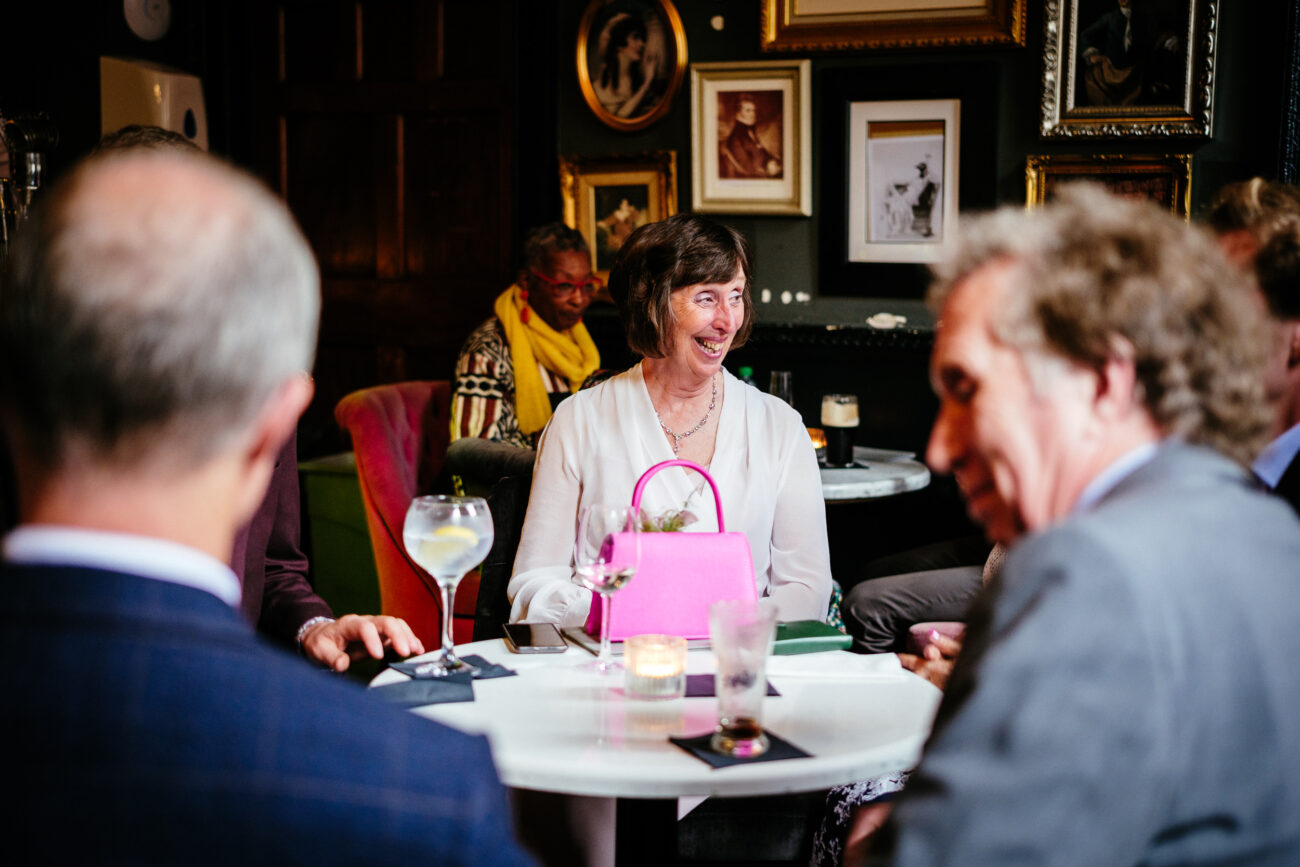 A group of people sit around a table in an art-filled room, engaging in conversation. A woman with a pink handbag smiles while others drink from various glasses.