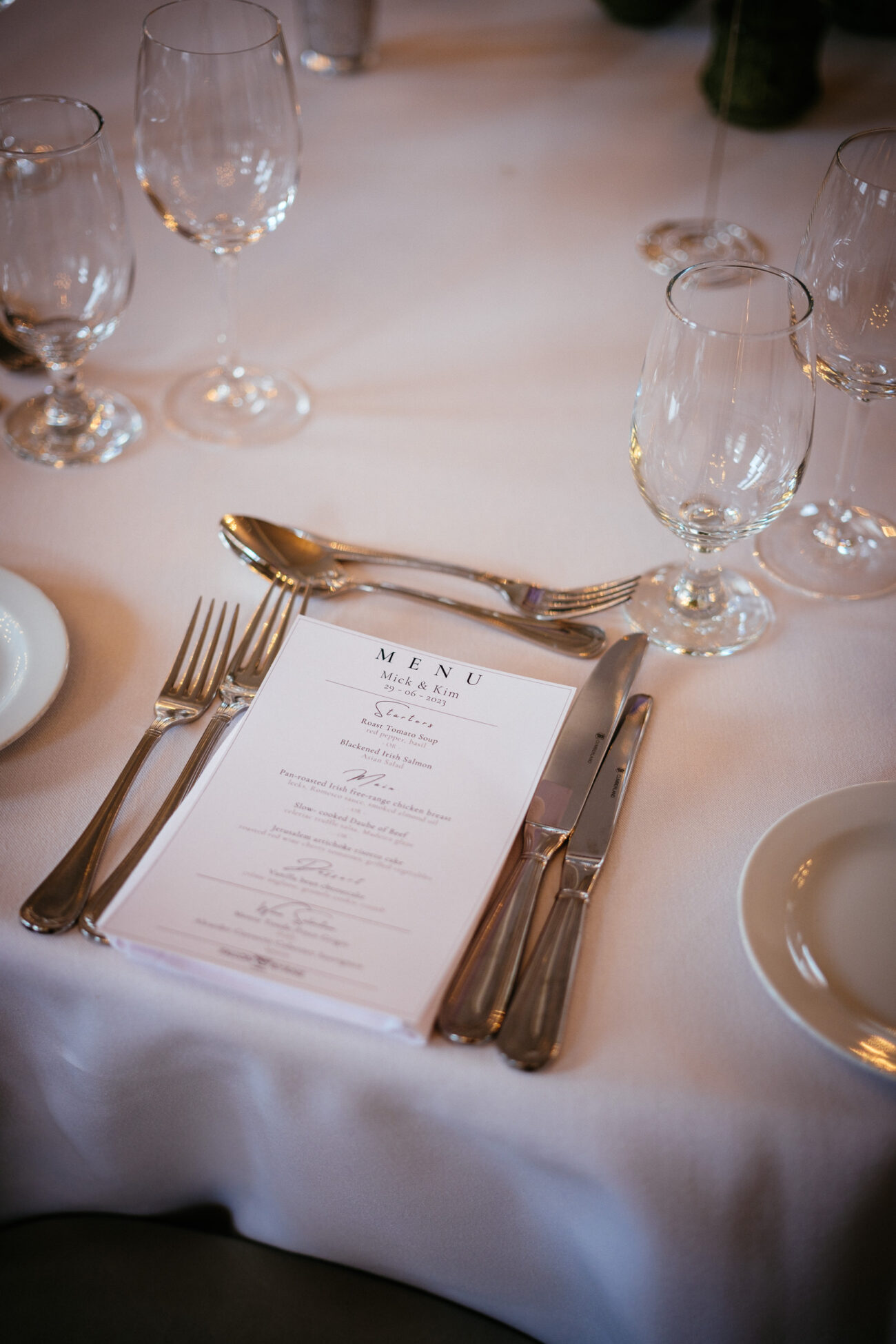 A table setting with a menu placed between a fork and knife on a napkin. Several wine and water glasses surround the setting on a white tablecloth.