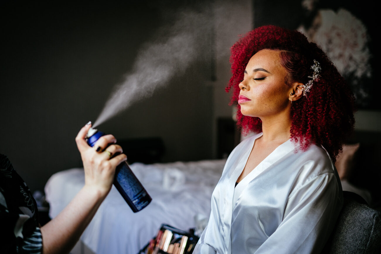 A person with red curly hair and a white robe has their eyes closed while another individual sprays hair spray onto their hair. The scene appears to be in a bedroom with makeup tools nearby.