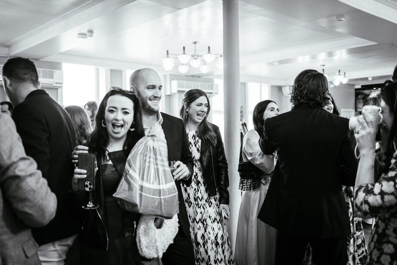 A group of people socializing at a lively indoor event, some holding drinks and shopping bags, with a modern chandelier overhead.