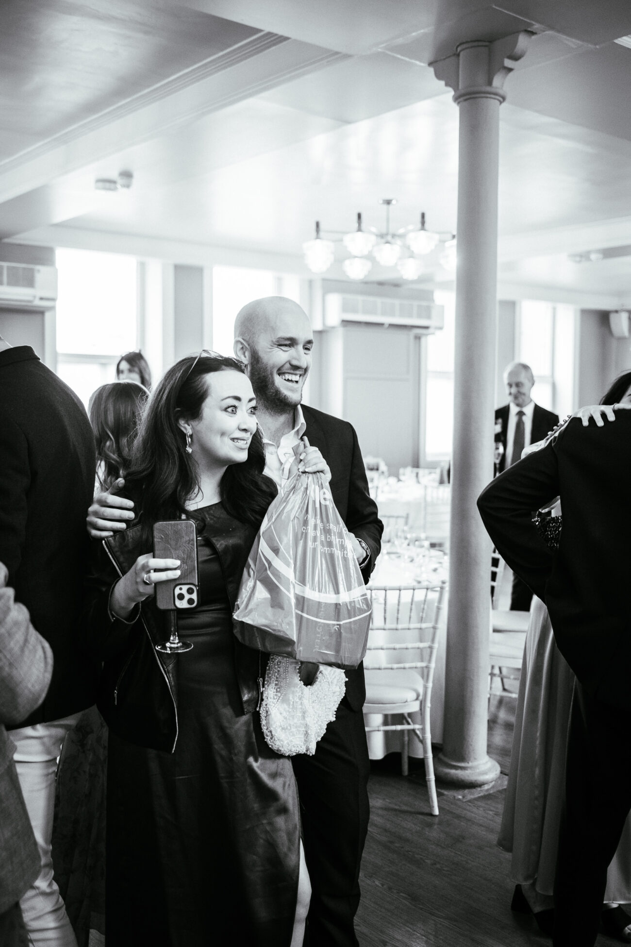 A man and woman, standing close to each other and smiling, are in a room with tables and chairs set up. The woman holds a drink and the man holds a plastic bag. Other people are present in the background.