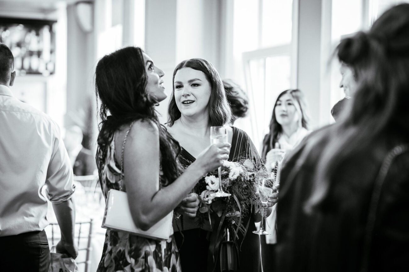A group of people engaging in conversation at a social event; one woman is holding a bouquet and another is holding a drink. The atmosphere is casual.