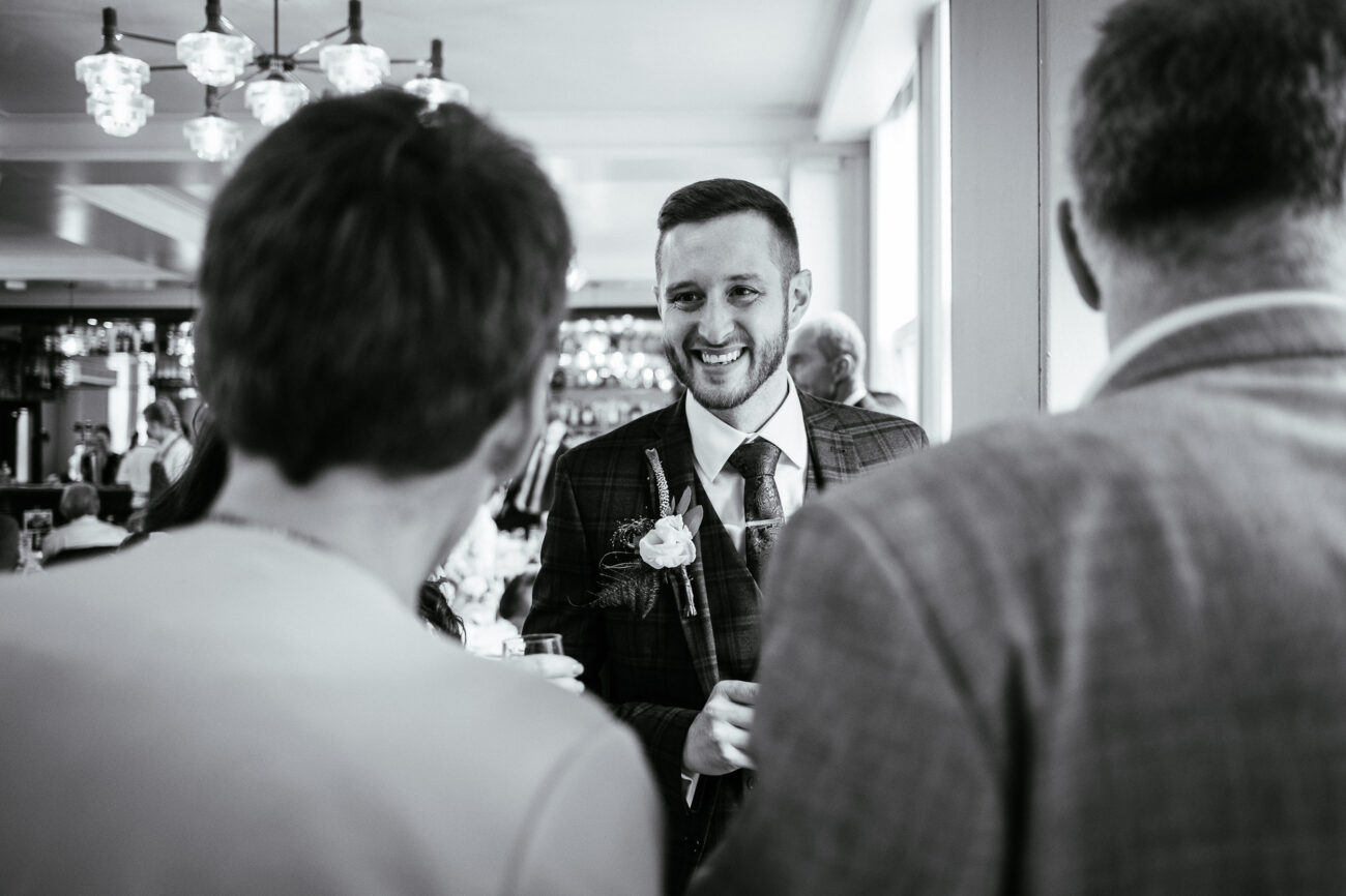 A man in a suit with a boutonniere smiles while talking to two other people in a well-lit indoor setting.