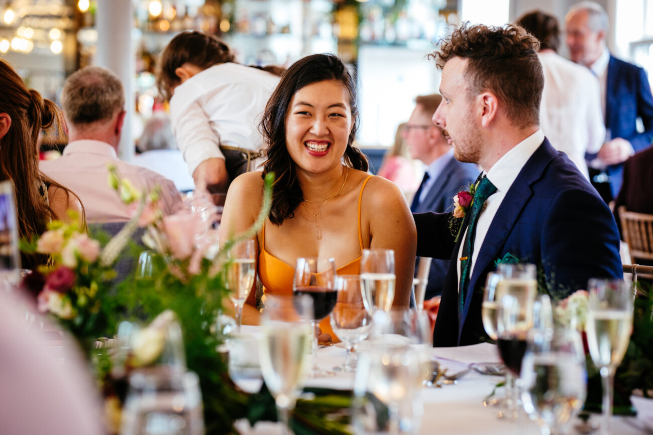A woman in an orange dress and a man in a suit smile while seated at a dining table with flowers and drinks at an event.
