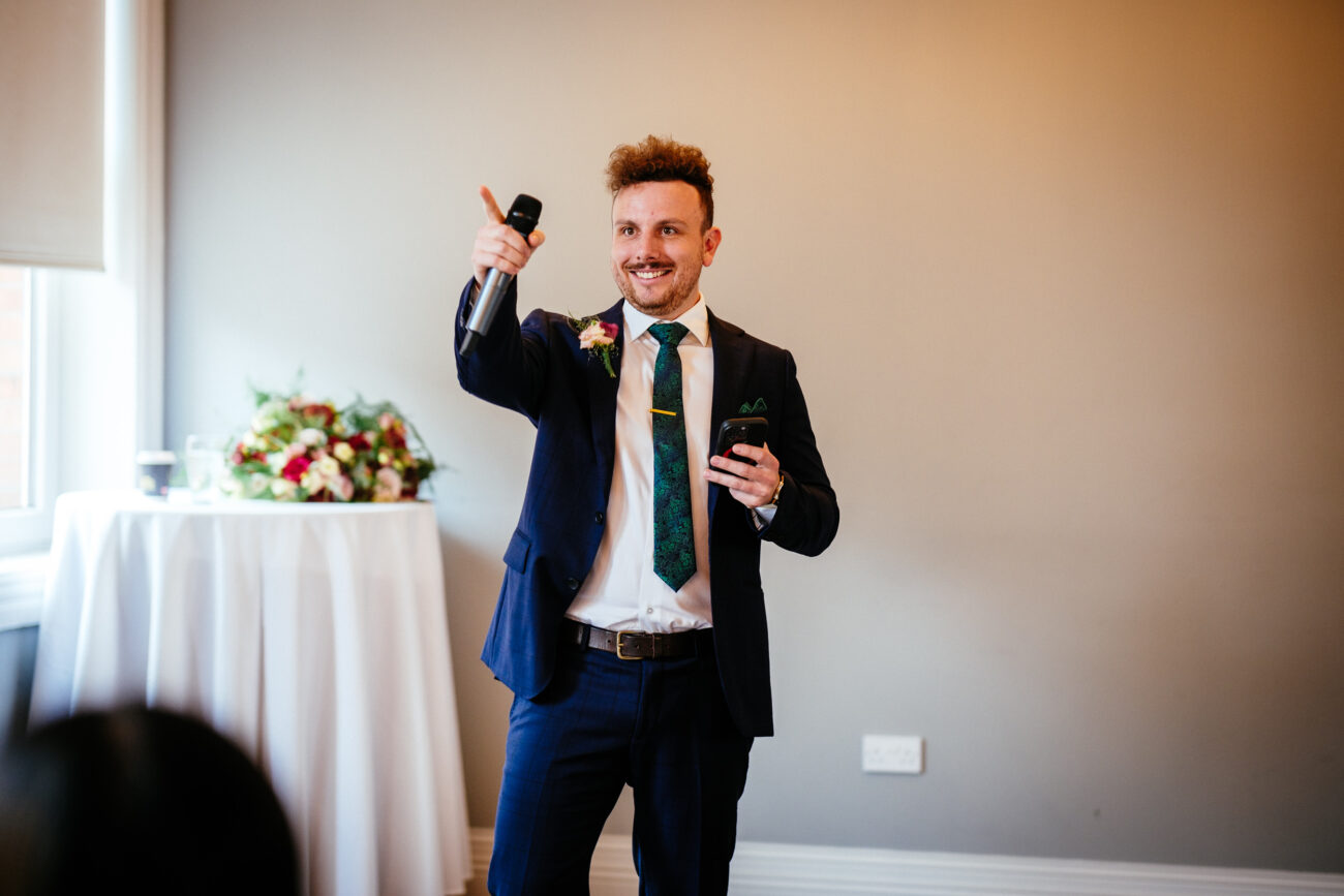A man in a blue suit and green tie holds a microphone and gestures while speaking. He stands near a table with a flower arrangement in a well-lit room.