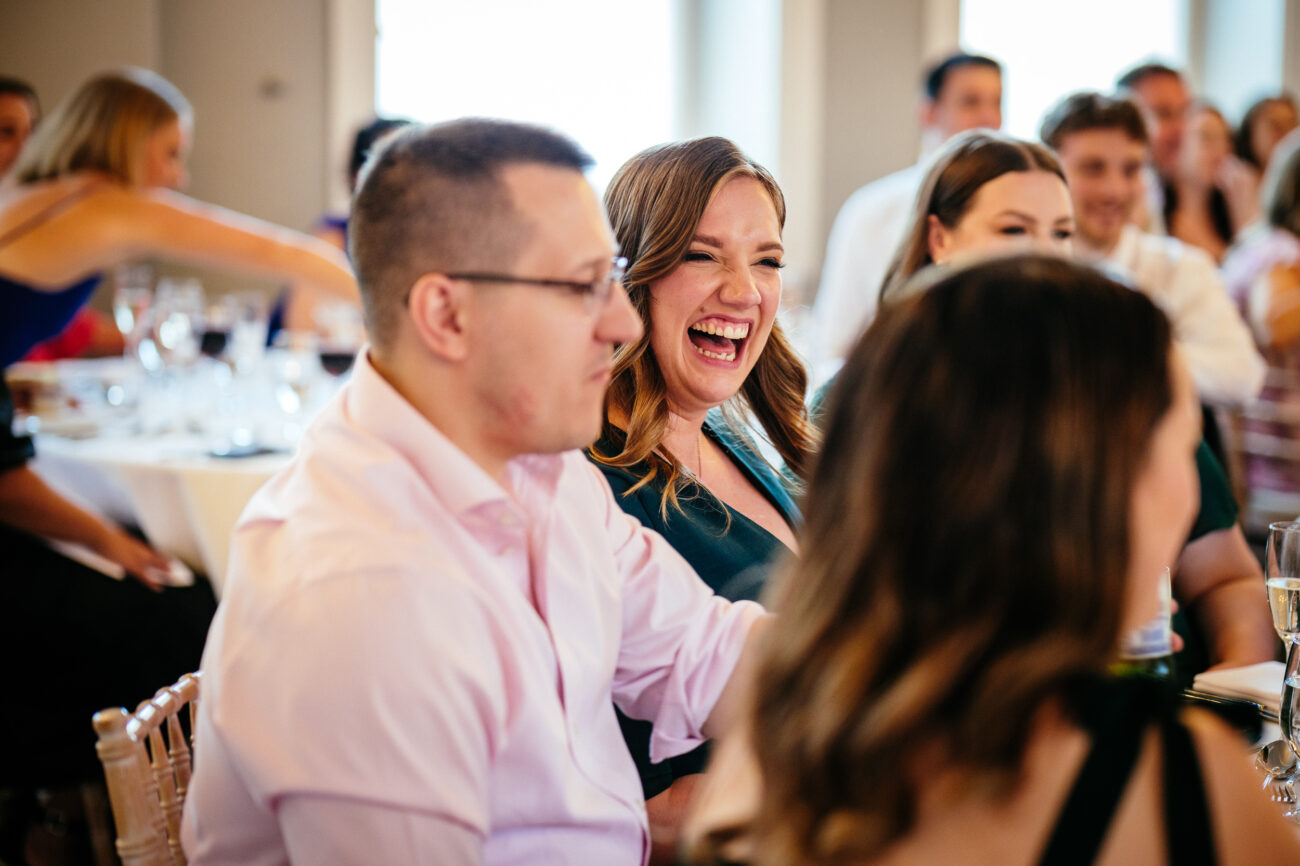 A group of people, seated at tables with white tablecloths and glassware, attentively listen while a woman in the foreground laughs heartily.
