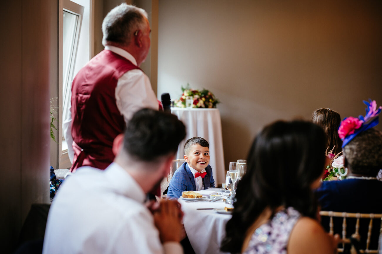 A smiling boy in a blue suit listens attentively to a man in a red vest standing and speaking in a room, with others seated around a table.