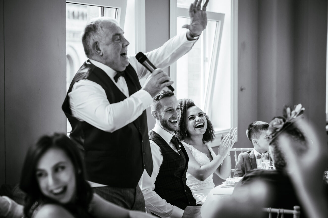 An elderly man with a microphone raises his hand while speaking at a wedding reception. A bride and groom seated next to him smile and clap. Guests at nearby tables are also visible in the background.