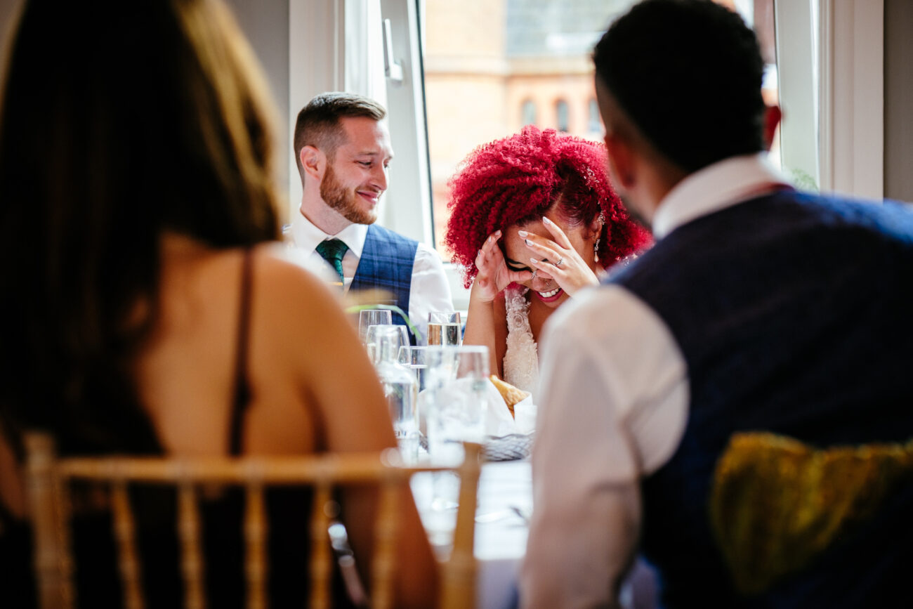 A woman with bright red hair covers her face while laughing, seated at a table with three other people, in a well-lit room with a window in the background.