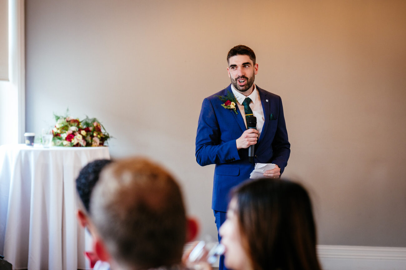 A man in a blue suit holding a microphone speaks to an audience at an indoor event. A flower arrangement is visible on a table near him.